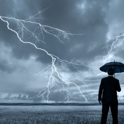 A rear view of a businessman in a suit, with an upheld umbrella, standing in a large field during a thunderstorm. Lightning is seen descending from a gray and cloudy sky.